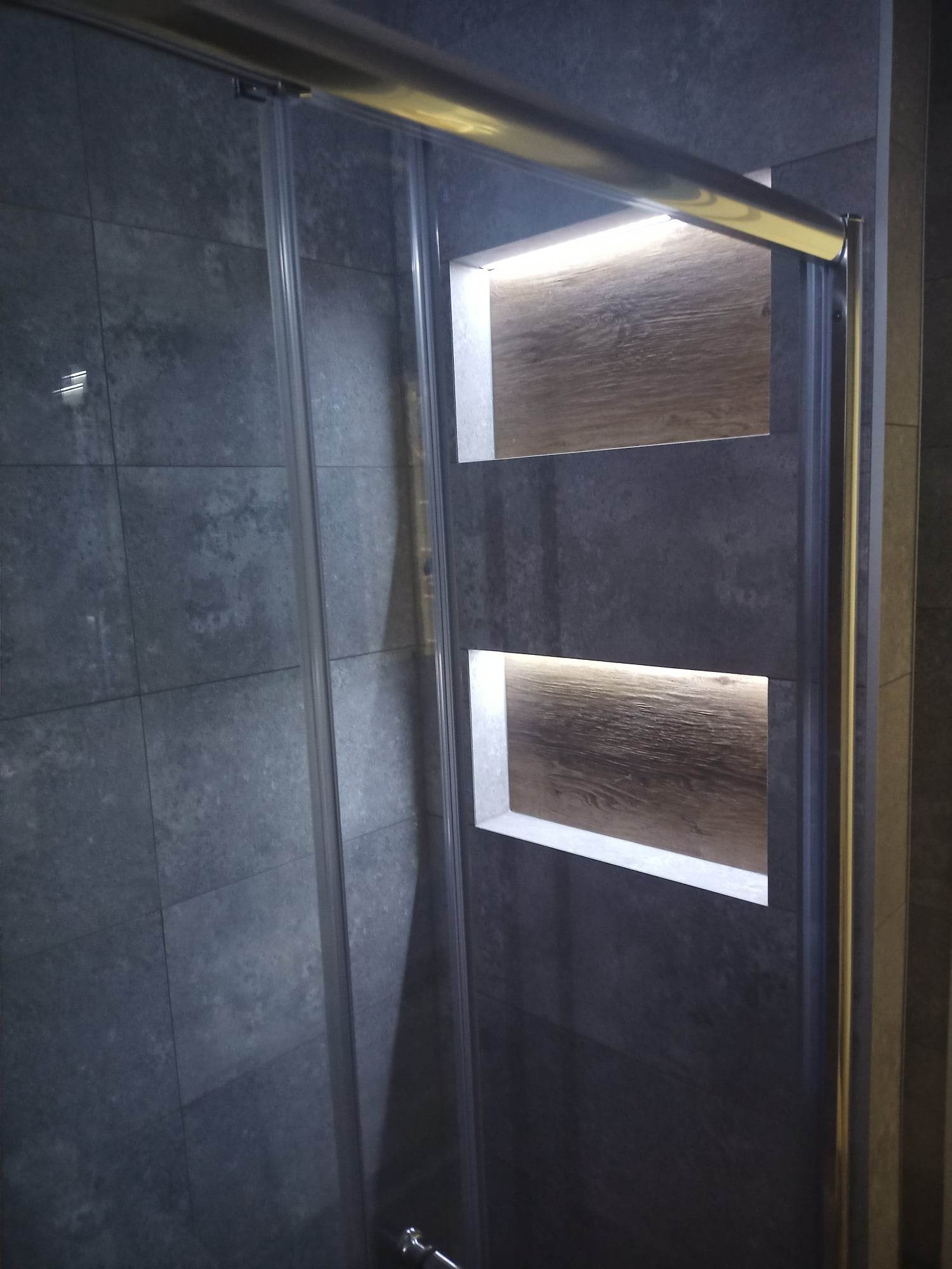 Kabina prysznicowa z przesuwnymi drzwiami, wykończona szarą, strukturalną płytką i wnękami z podświetleniem LED, imitującymi drewno.