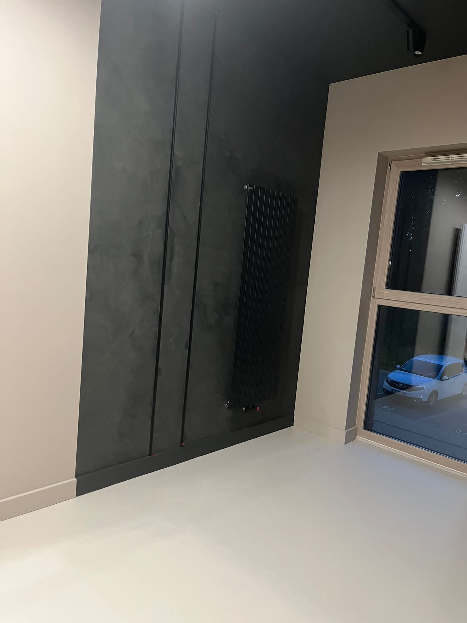Nowoczesne wnętrze z ciemnoszarą ścianą z dekoracyjnymi pionowymi wstawkami, pionowym grzejnikiem i jasną podłogą, widok na zaparkowany samochód za oknem.