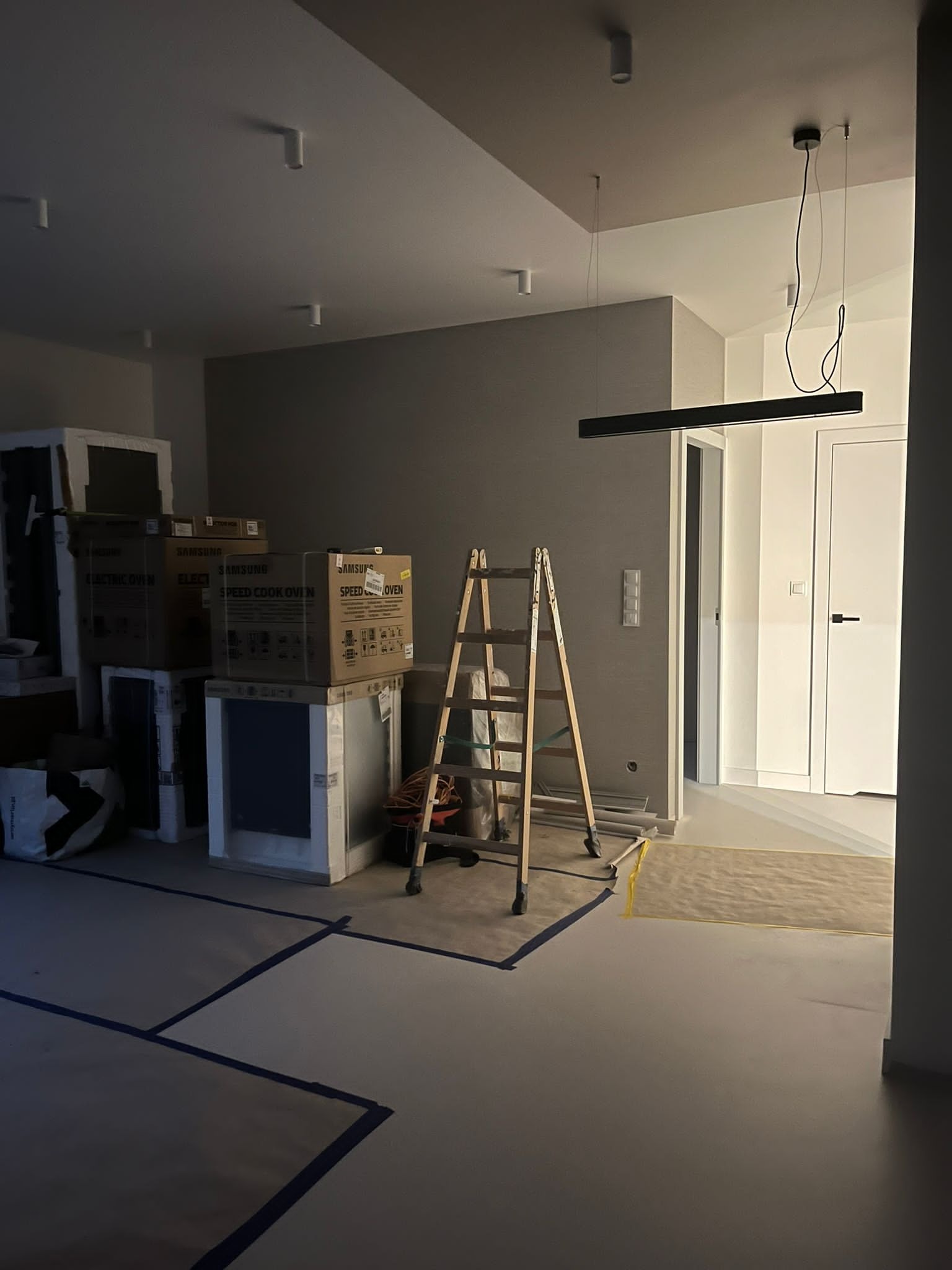 Wnętrze w trakcie prac wykończeniowych: kartony z nowymi piekarnikami Samsung, drabina, zabezpieczona podłoga z wyznaczonymi obszarami, nowoczesne oświetlenie sufitowe.