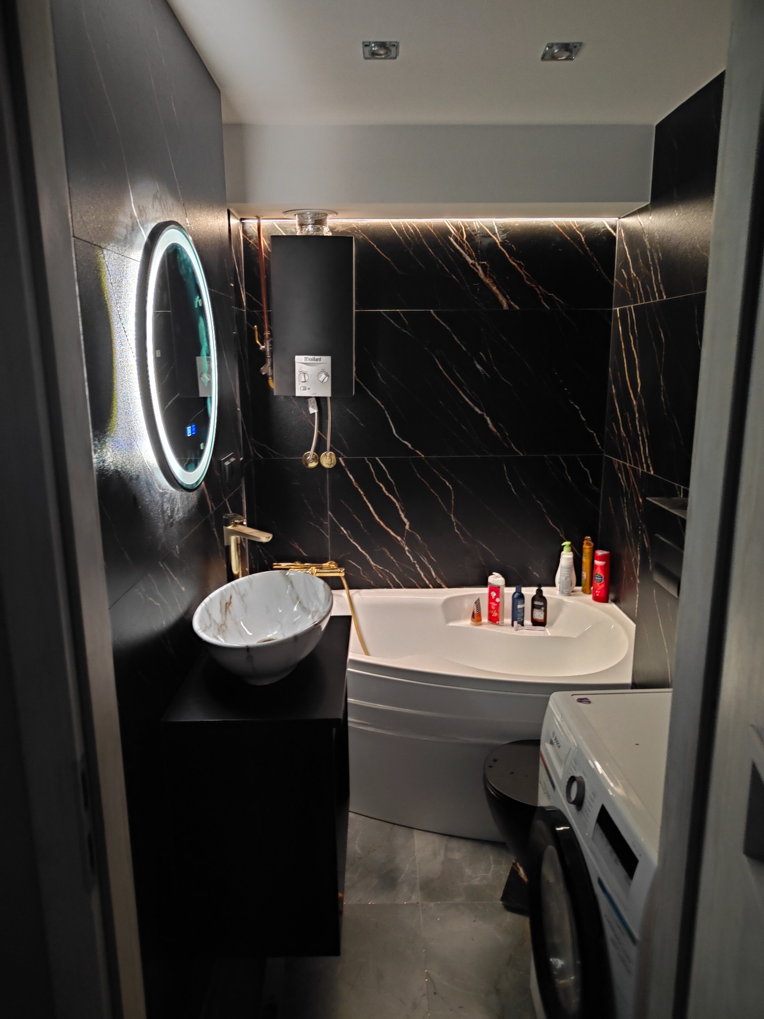 Nowoczesna łazienka z czarnymi płytkami imitującymi marmur, okrągłym lustrem z podświetleniem LED, owalną umywalką nablatową, wanną narożną i pralką.