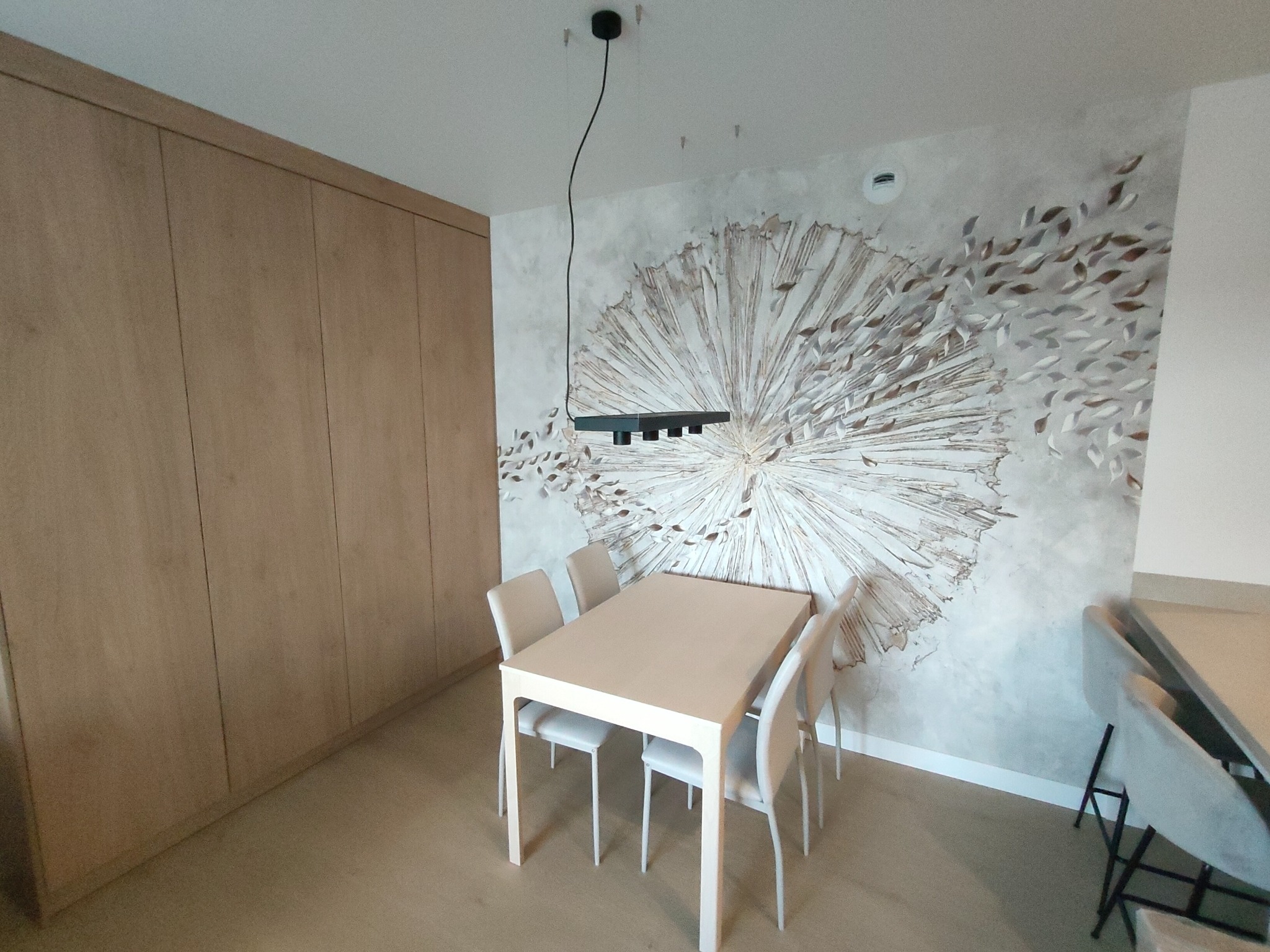 Jasne, minimalistyczne wnętrze jadalni z drewnianą zabudową ścienną, stołem z krzesłami o jasnej kolorystyce oraz dekoracyjną tapetą z motywem rozchodzących się liści na ścianie. Nad stołem wisi...