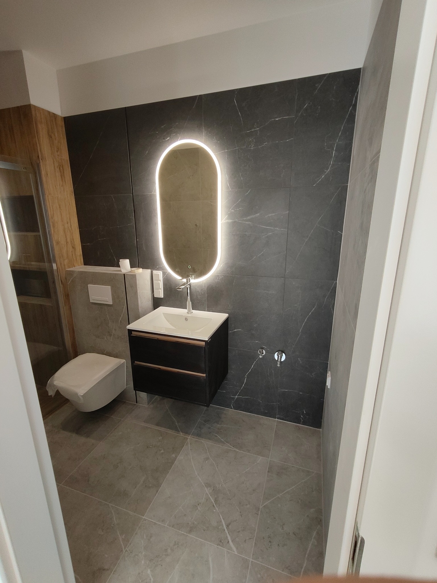 Nowoczesna łazienka z szarymi płytkami imitującymi marmur na ścianach i podłodze, podświetlane owalne lustro nad czarną szafką umywalkową, wisząca toaleta z zabudowanym systemem spłukiwania.