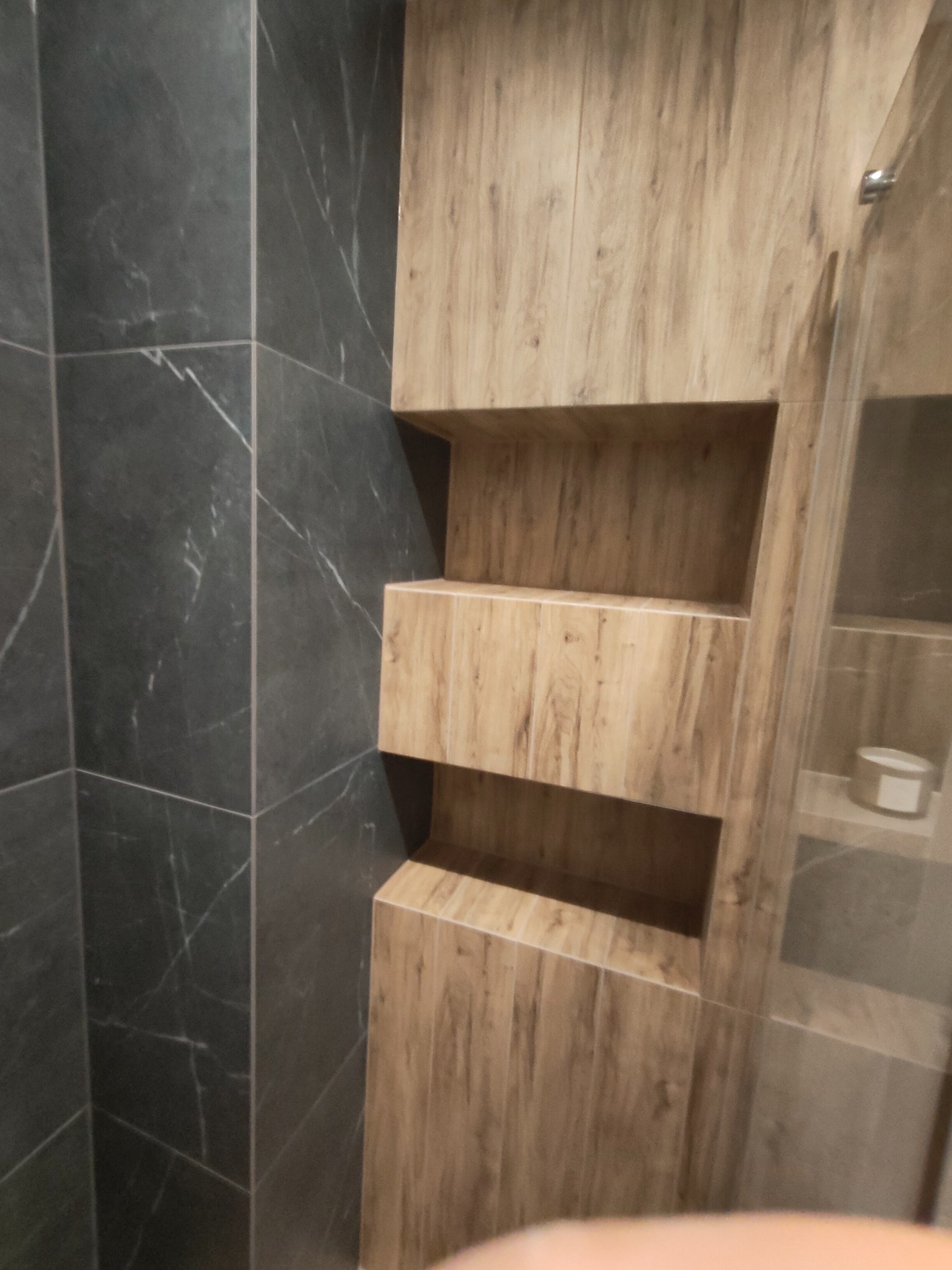 Wykończona wnęka z półkami z drewnopodobnych paneli w łazience z ciemnymi, marmurkowymi płytkami.
