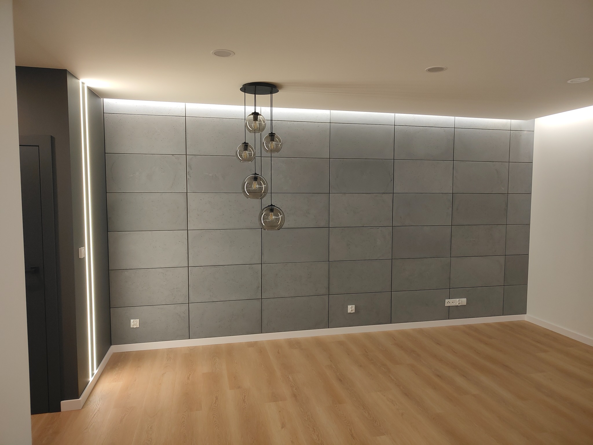 Nowoczesne wnętrze z drewnianą podłogą, ścianą wyłożoną szarymi płytami imitującymi beton, oświetleniem LED wzdłuż sufitu i wiszącą lampą z szklanymi kulami.