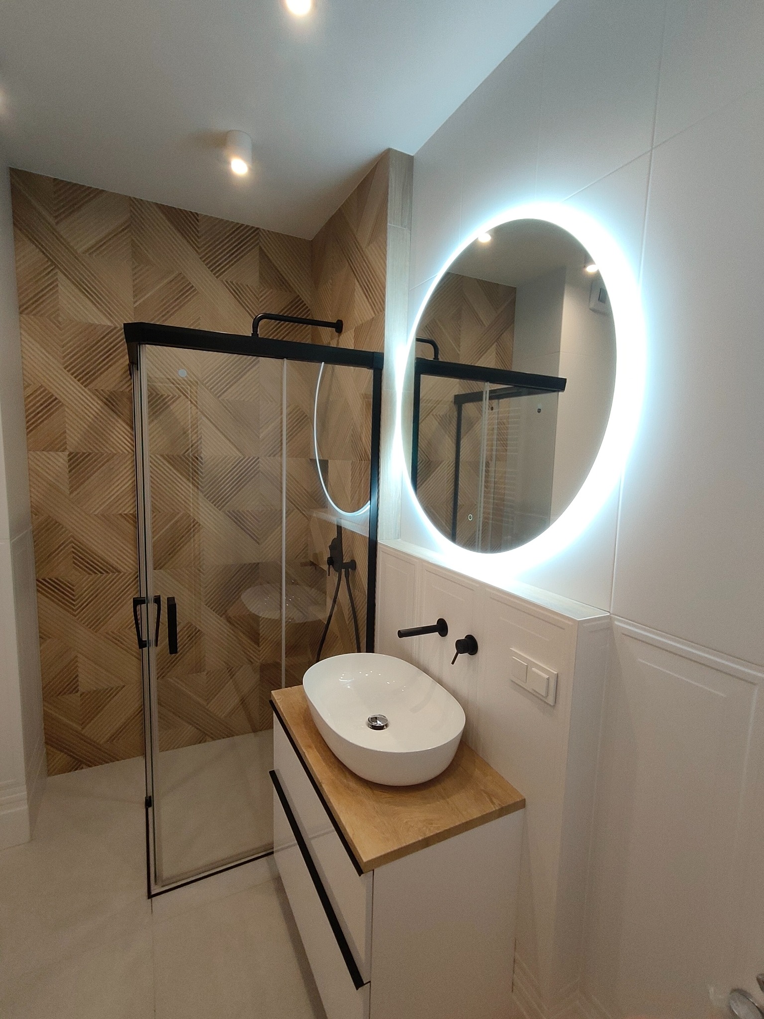Nowoczesna łazienka z kabiną prysznicową z czarnymi ramami, umywalką nablatową na szafce z drewnianym blatem, okrągłym lustrem z podświetleniem LED i ścianą wyłożoną płytkami imitującymi drewno.
