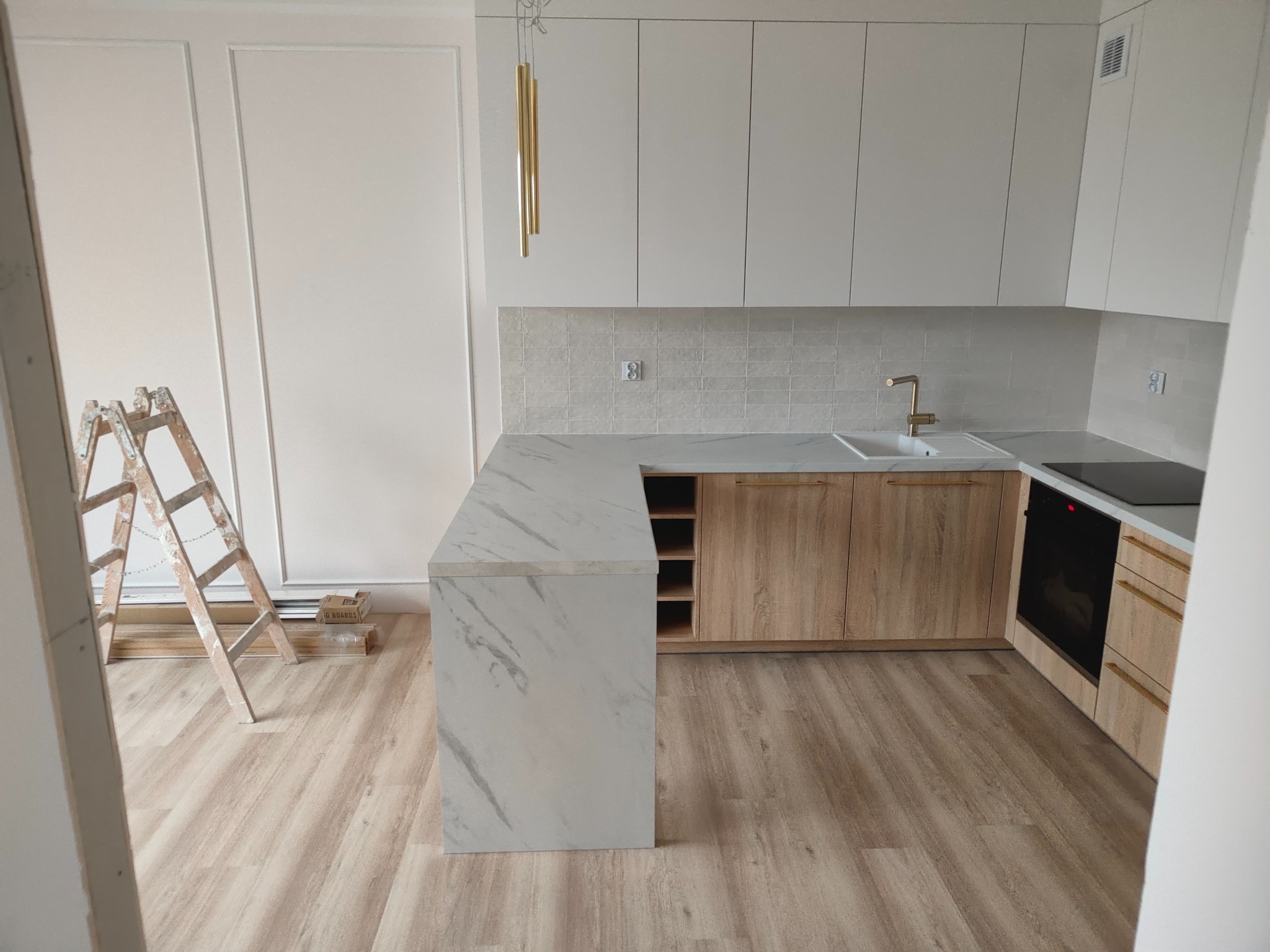 Wnętrze jasnej kuchni w trakcie wykańczania, widoczna drabina oparta o ścianę z dekoracyjnymi listwami, marmurowy blat, drewniane szafki i panele podłogowe ułożone w jodełkę.