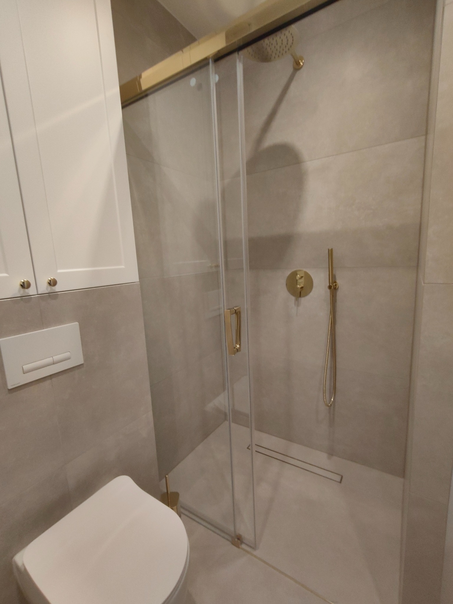 Nowoczesna kabina prysznicowa z przesuwnymi drzwiami, złotymi detalami i odpływem liniowym, obok toaleta i szafka z białymi frontami.