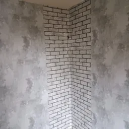 Fragment pokoju z wykończonym narożnikiem: ściana pokryta szarą strukturą dekoracyjną sąsiaduje z pionowym pasem tapety imitującej białą cegłę z szarą fugą.