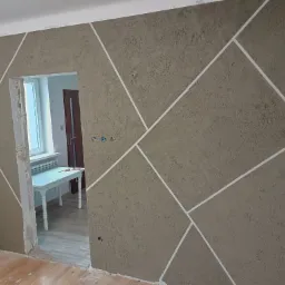 Ściana pokryta strukturalną gładzią w odcieniach brązu, zdobiona białymi, geometrycznymi liniami tworzącymi asymetryczny wzór, widok na remontowane wnętrze z drabiną i stołem.