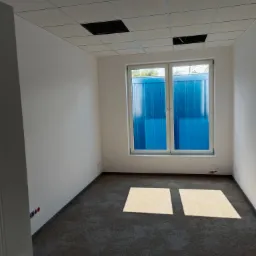 Pusty pokój biurowy z szarym dywanem, białymi ścianami i kasetonowym sufitem z brakującymi panelami, widok na błękitny budynek przez okno.