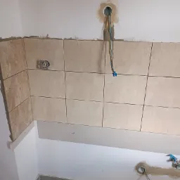 Ściana w trakcie układania płytek ceramicznych w kuchni, widoczne puszki elektryczne i wystające przewody, częściowo położona płyta karton gips, wiadro farby na podłodze.