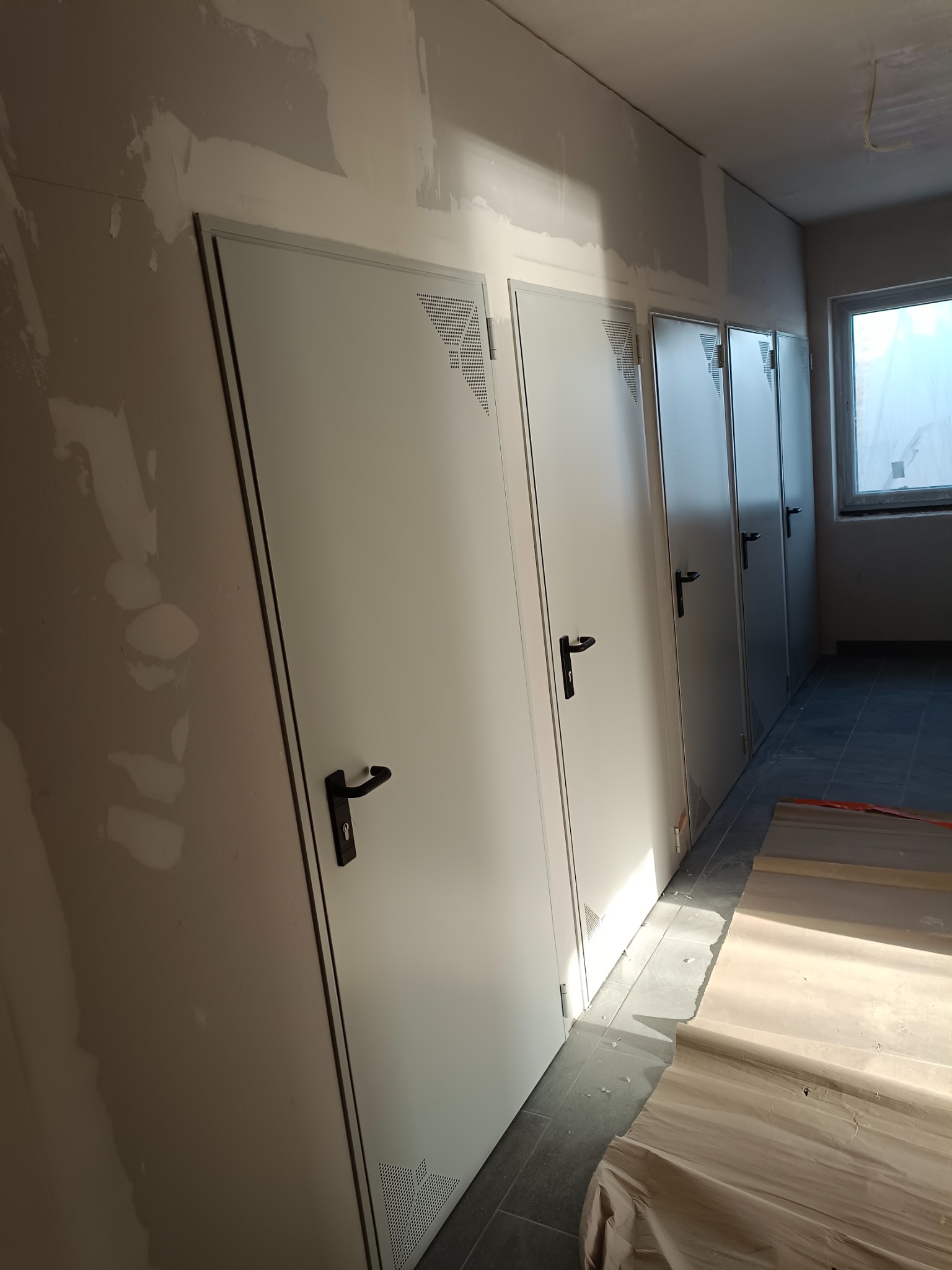 Ściana w trakcie wykańczania, widoczne ślady po szpachlowaniu, rząd białych drzwi z czarnymi klamkami, na podłodze folia ochronna.