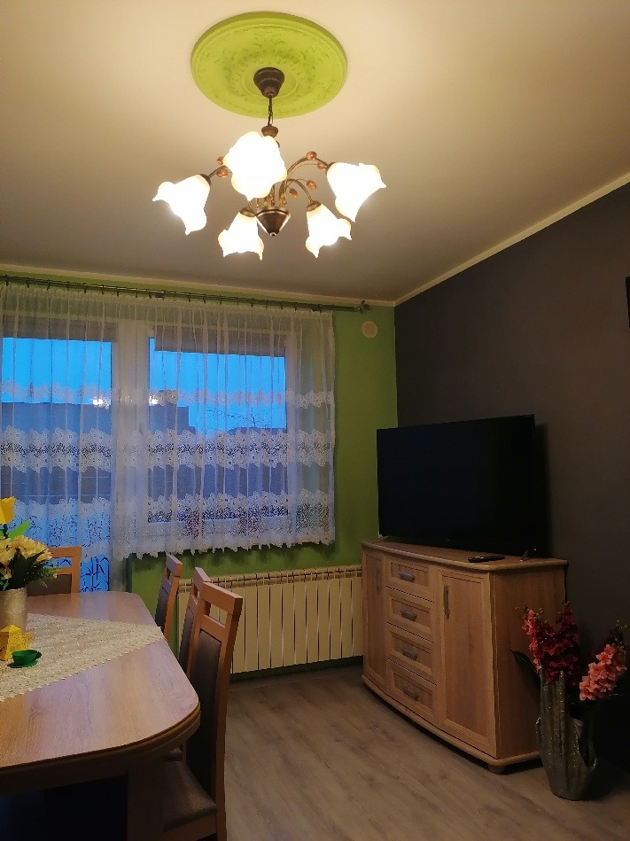 Jadalnia z drewnianym stołem, krzesłami, komodą, telewizorem i dekoracyjnym żyrandolem z zielonym rozetą sufitową.