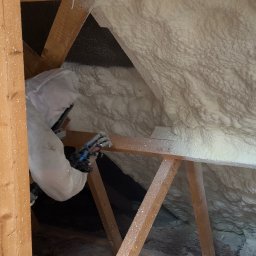 Paweł Żuchowski TERMO-DOM - Pracownik w kombinezonie ochronnym aplikuje pianę izolacyjną w przestrzeni poddasza w Szczecinie, widoczne drewniane elementy konstrukcyjne dachu.