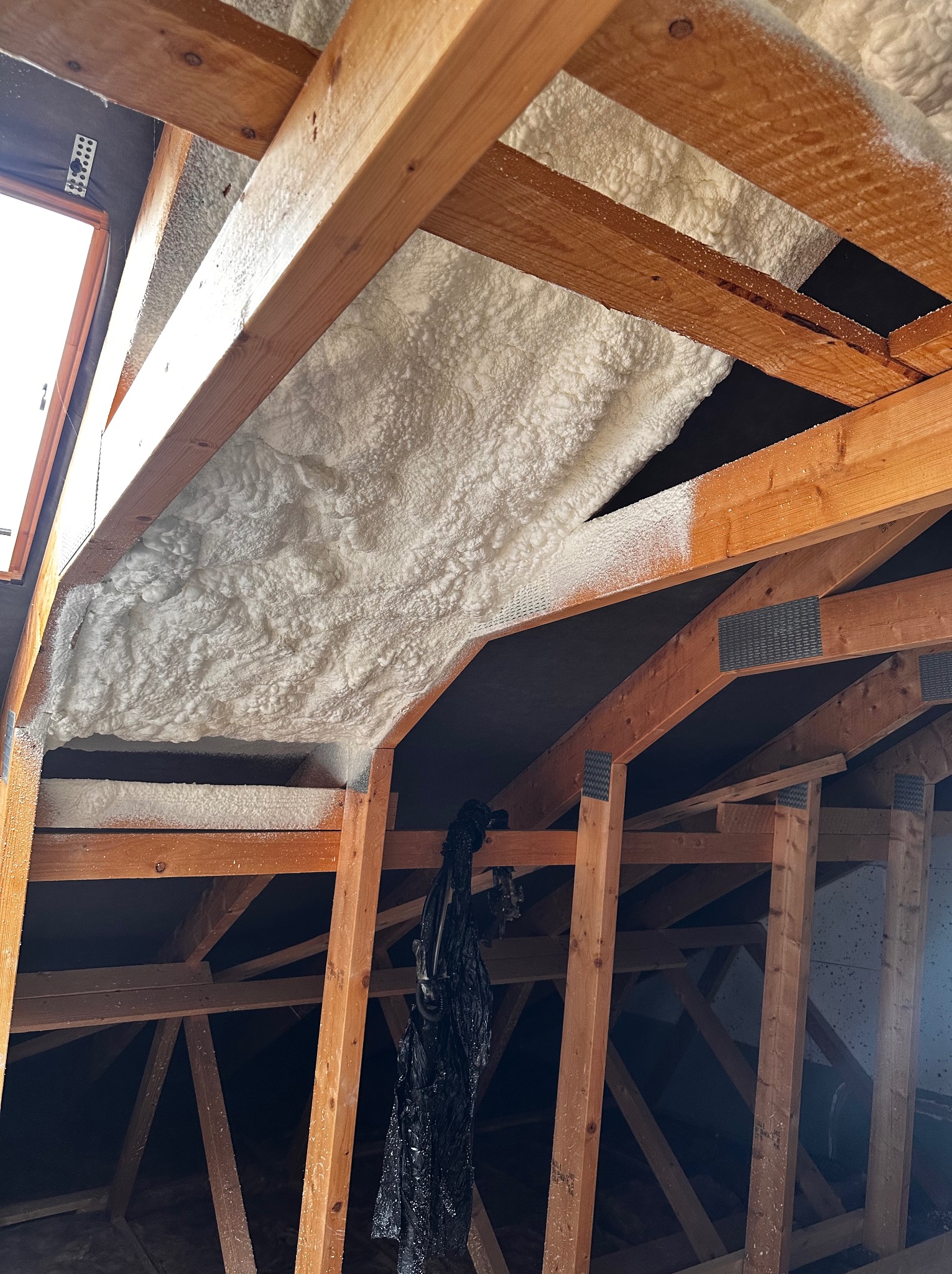 Poddasze z drewnianą konstrukcją krokwiową, częściowo pokryte białą, grubą warstwą izolacji natryskowej, widoczne elementy mocujące i fragment okna dachowego.