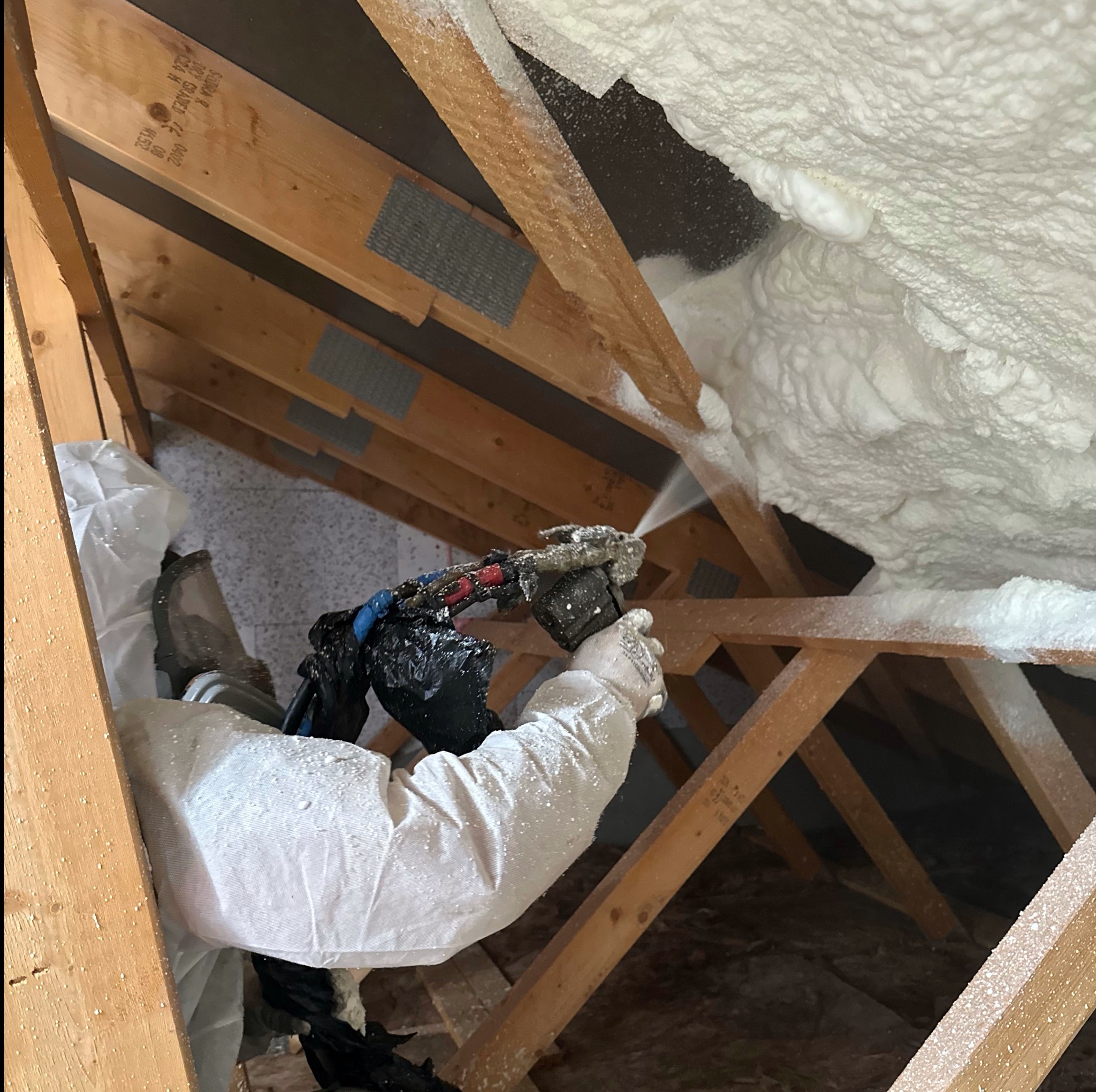 Pracownik w kombinezonie ochronnym aplikuje pianę izolacyjną na drewnianą konstrukcję poddasza, widoczne kropelki piany w powietrzu i świeżo nałożona warstwa izolacji.