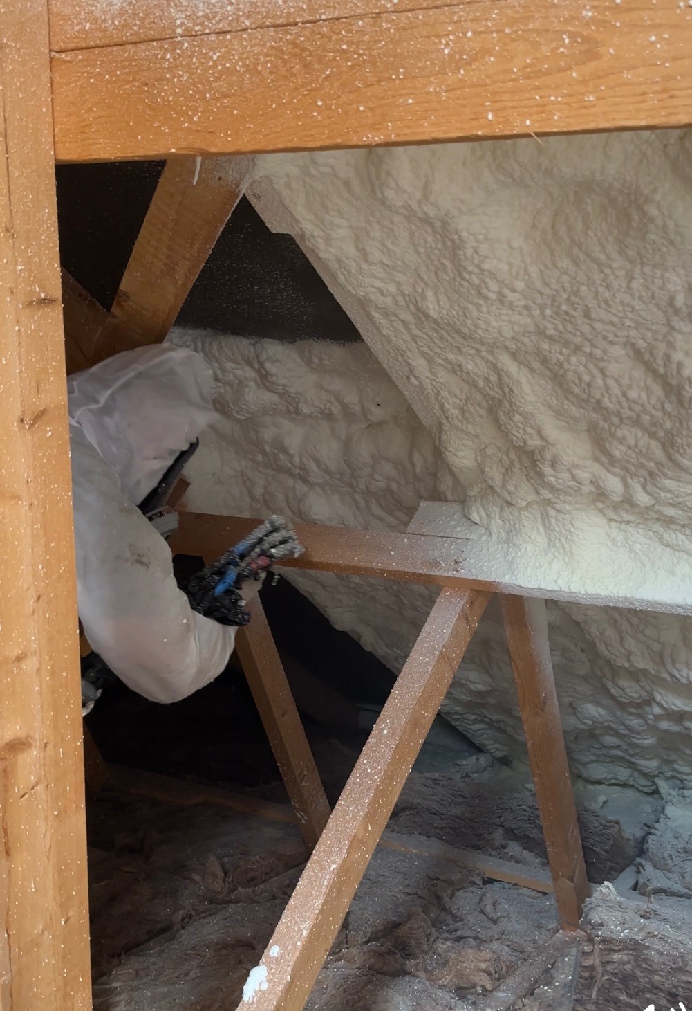 Pracownik w kombinezonie ochronnym aplikuje pianę izolacyjną w przestrzeni poddasza w Szczecinie, widoczne drewniane elementy konstrukcyjne dachu.