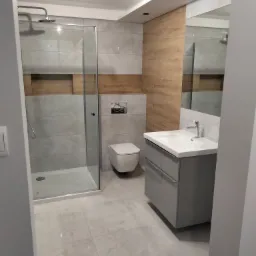 Nowoczesna łazienka z kabiną prysznicową ze szklanymi drzwiami, podwieszaną toaletą, szarą szafką z umywalką i drewnopodobnymi płytkami na ścianie, oświetlona reflektorami.