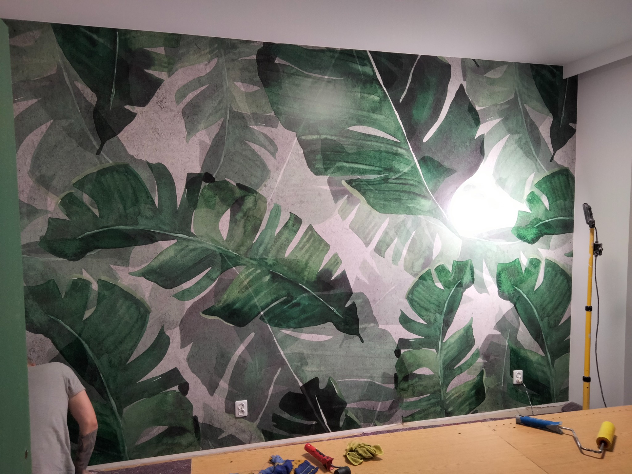 Ściana pokryta tapetą z motywem tropikalnych liści, widoczne narzędzia malarskie i fragment sylwetki osoby w trakcie pracy.