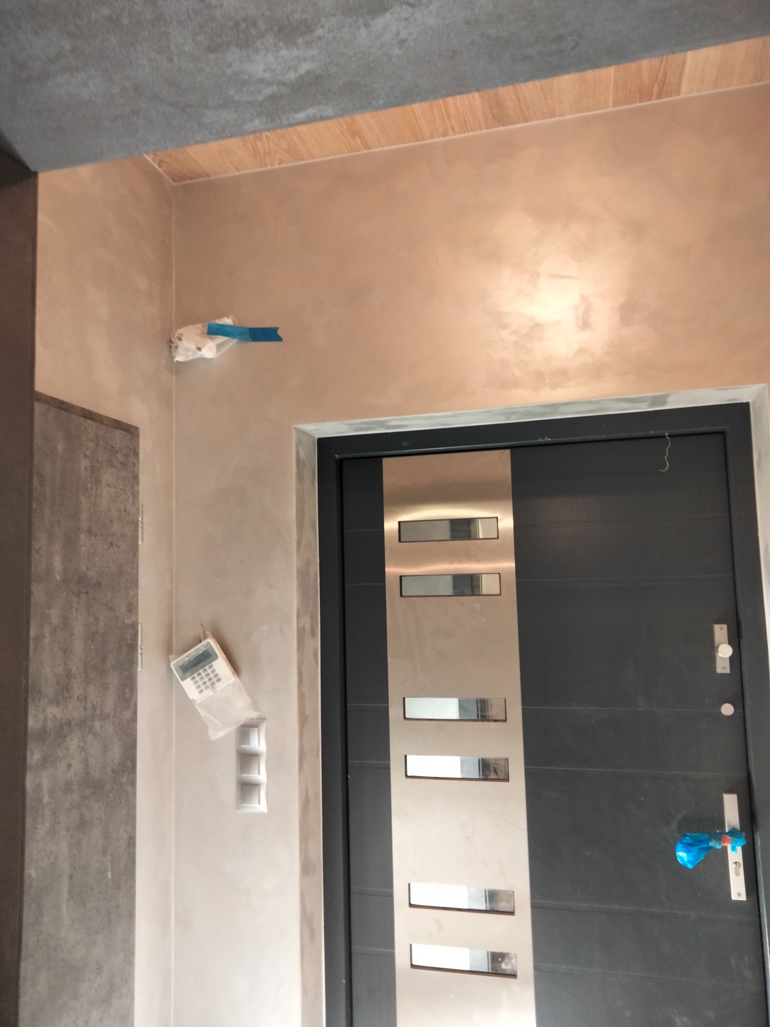 Ściana w przedpokoju świeżo po malowaniu dekoracyjnym w odcieniach beżu i brązu, zabezpieczone elementy instalacji elektrycznej i klamka drzwi wejściowych folią malarską, widoczne włączniki światła...