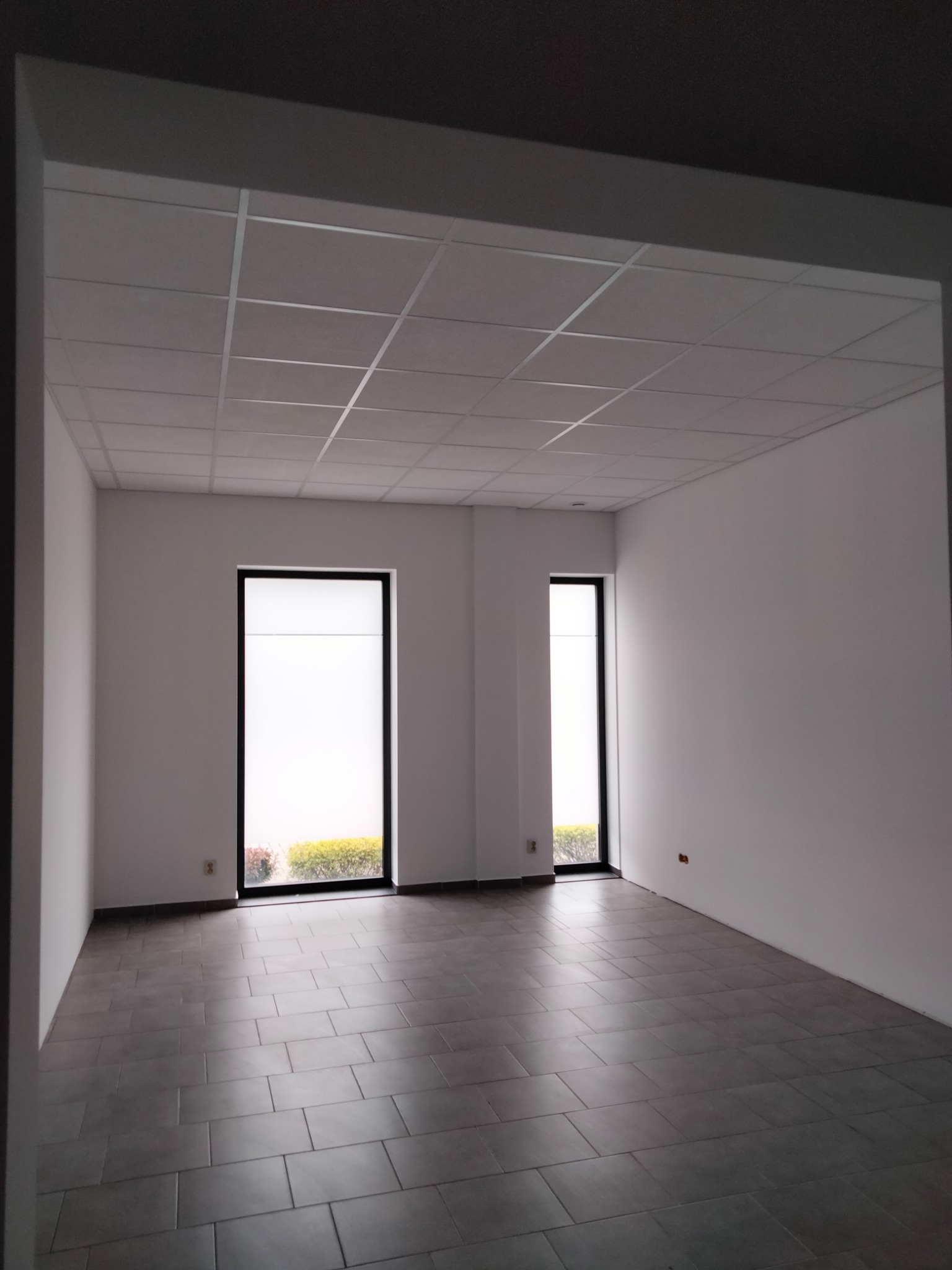 Puste, odnowione pomieszczenie biurowe z szarą podłogą z płytek, białymi ścianami, podwieszanym sufitem kasetonowym i dwoma oknami z widokiem na zieleń.