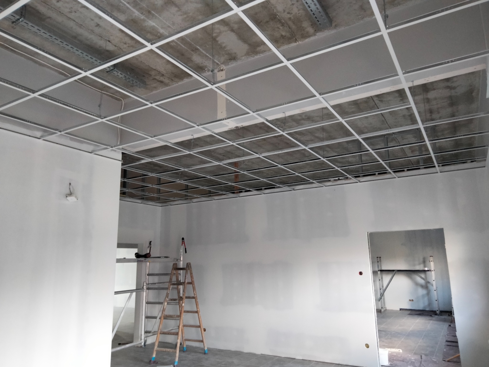 Montaż sufitu podwieszanego z płyt kartonowo-gipsowych, widoczna metalowa konstrukcja kratowa i częściowo zamontowane płyty, drabina w tle, surowy betonowy strop powyżej konstrukcji.