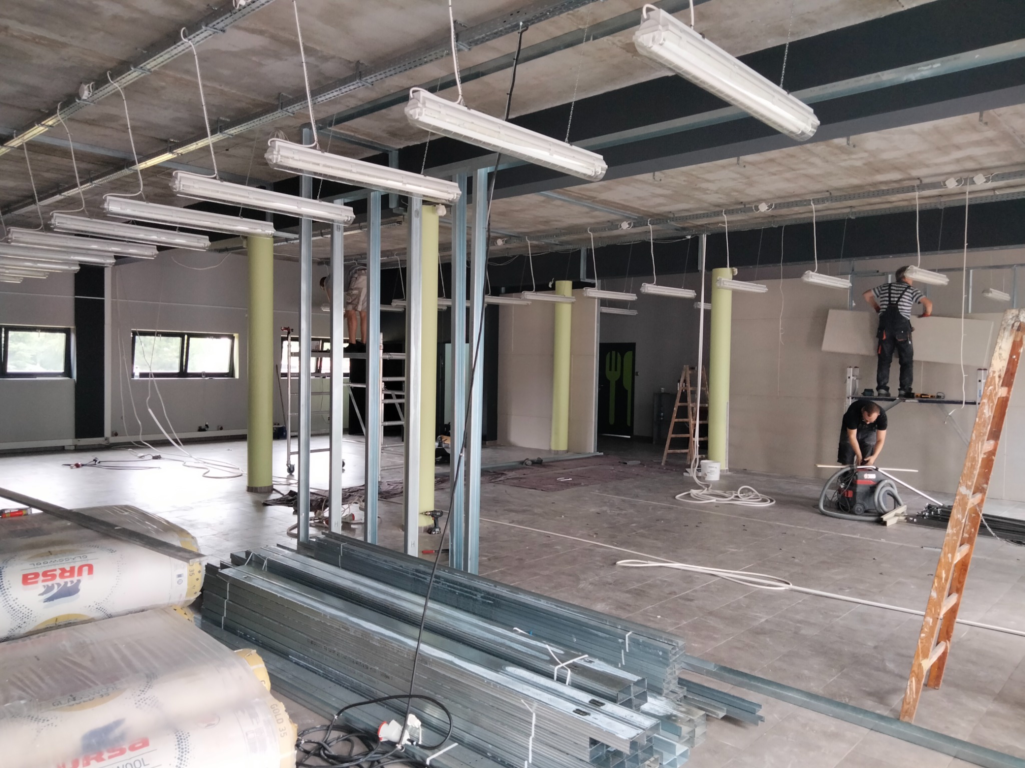 Wnętrze biura w trakcie remontu: montaż metalowych profili, położona izolacja, pracownicy na rusztowaniu montują płytę karton gips, widoczne narzędzia i odkurzacz przemysłowy.