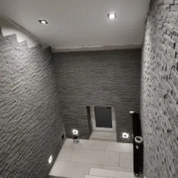 Korytarz w trakcie wykańczania z szarą okładziną imitującą kamień na ścianach, podświetlony punktowymi lampami w suficie i w ścianach, widoczne schody wyłożone płytkami oraz okno w ścianie, ślady...