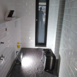 Profbud - Wnętrze łazienki w trakcie remontu z białymi, prostokątnymi płytkami na ścianach, zabezpieczoną podłogą, widocznymi instalacjami hydraulicznymi i czarną walizką narzędziową w rogu.