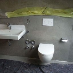 Profbud - Montaż umywalki i toalety w łazience z szarą okładziną, zabezpieczona podłoga, widoczne przygotowane instalacje.