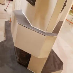 Wykończony kominek z szarymi płytkami i kremowymi panelami, widoczny fragment paleniska, w tle osoba sprzątająca podłogę.