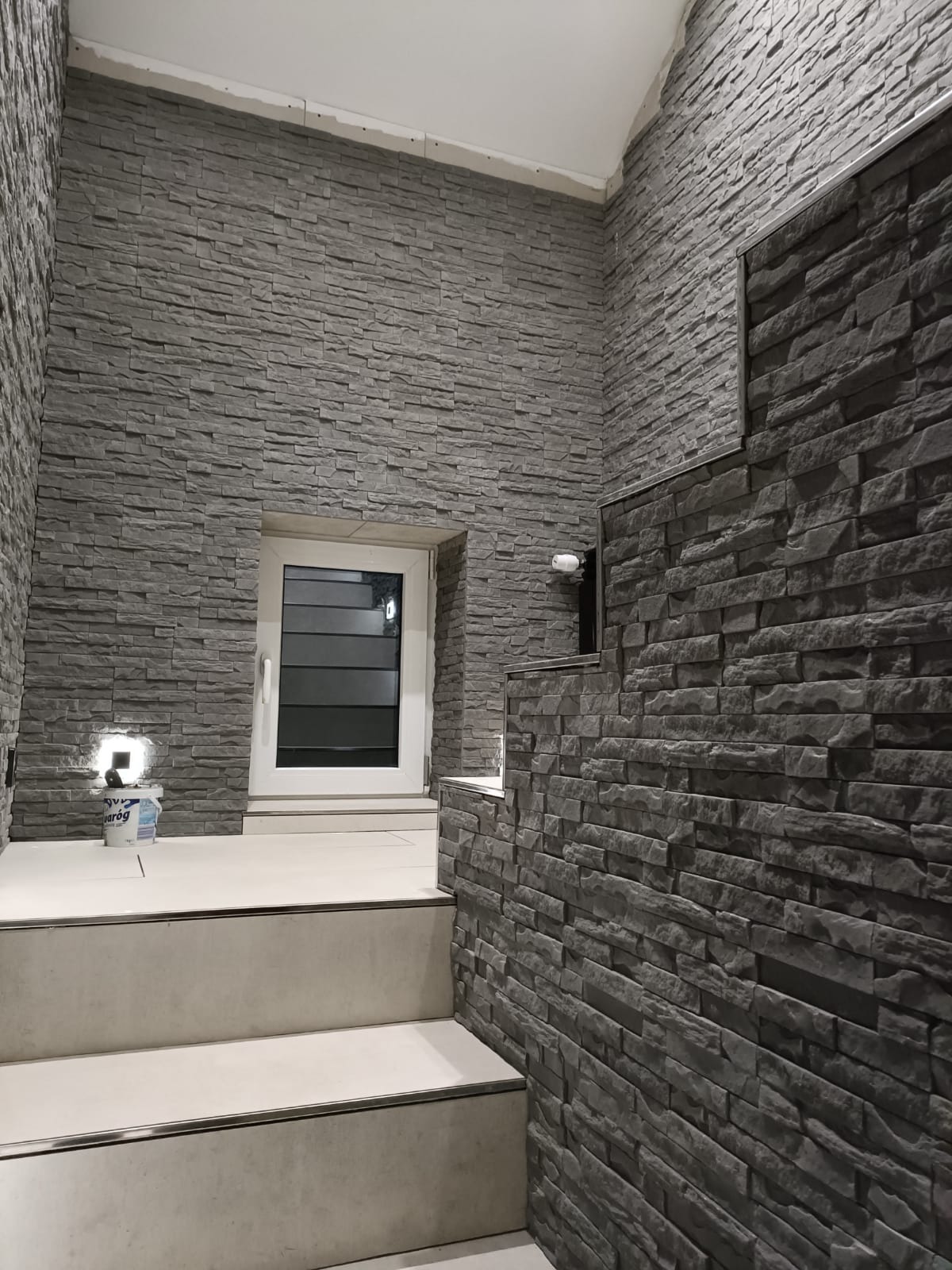 Korytarz wykończony szarą okładziną kamienną, widoczne schody z jasnymi płytkami i okno z żaluzjami. Na podeście stoi wiadro z farbą i lampka.