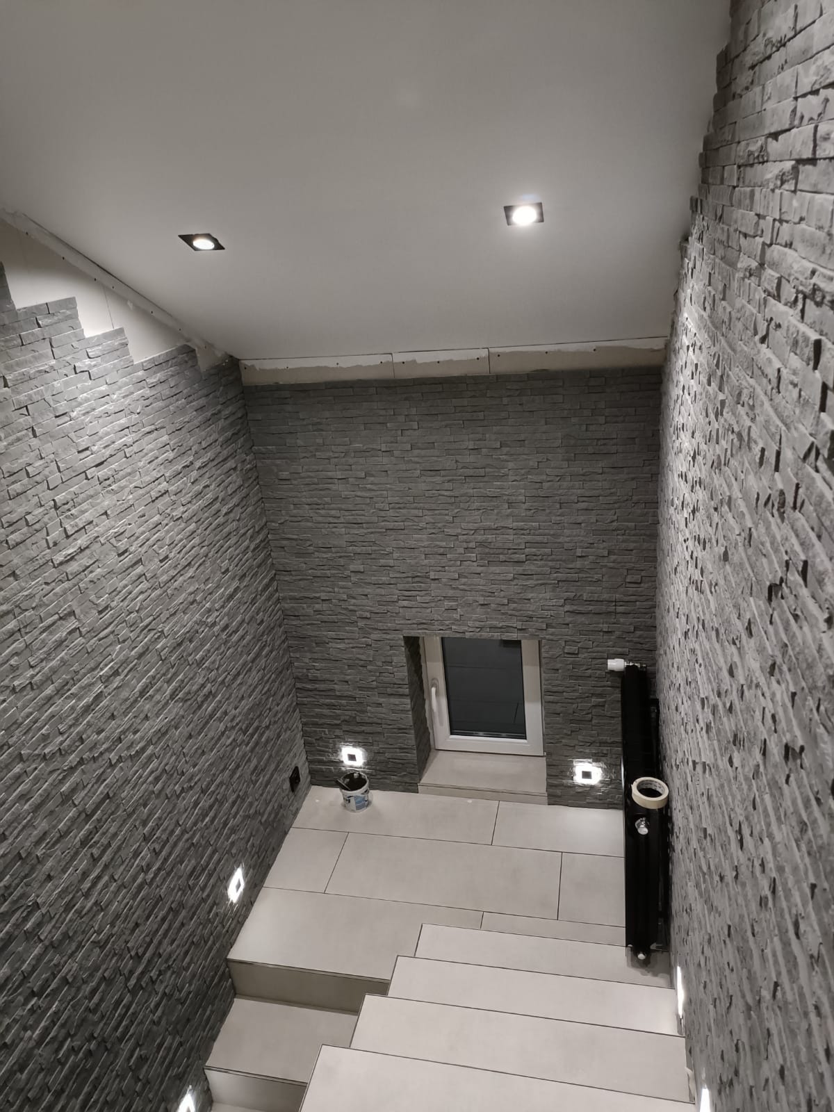 Korytarz w trakcie wykańczania z szarą okładziną imitującą kamień na ścianach, podświetlony punktowymi lampami w suficie i w ścianach, widoczne schody wyłożone płytkami oraz okno w ścianie, ślady...
