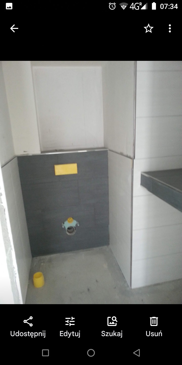 Wnętrze łazienki w trakcie remontu: szare płytki na ścianie z otworem na instalację, białe płytki na pozostałych ścianach, żółty element instalacyjny i kubek na podłodze.