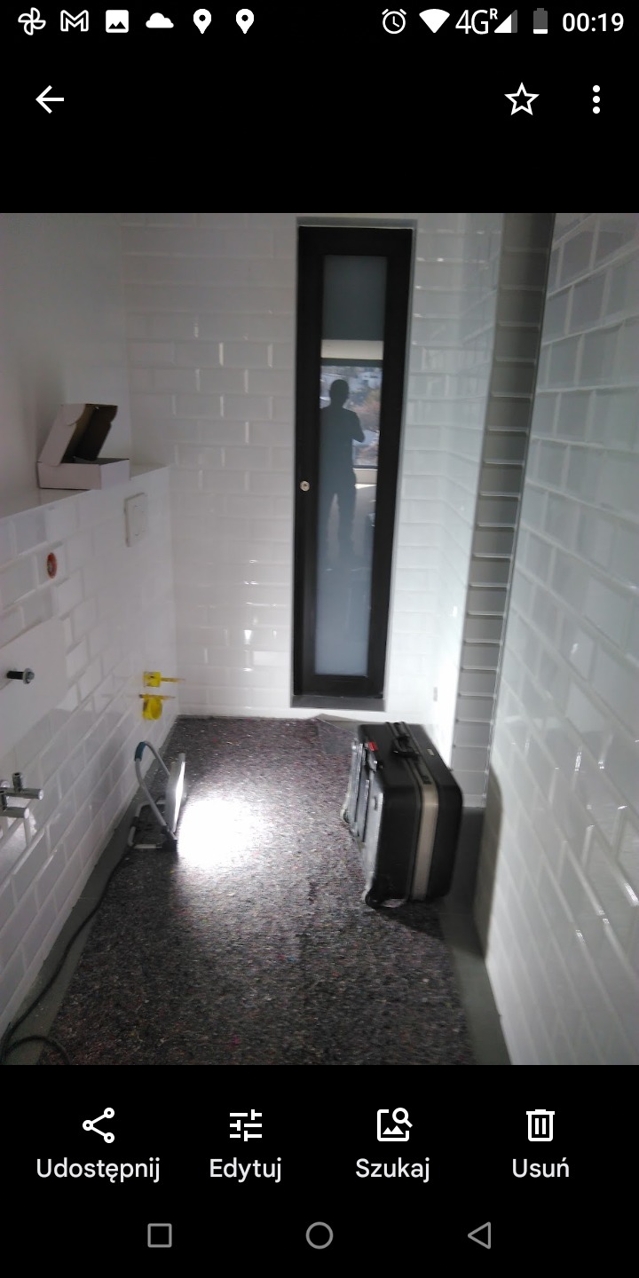 Wnętrze łazienki w trakcie remontu z białymi, prostokątnymi płytkami na ścianach, zabezpieczoną podłogą, widocznymi instalacjami hydraulicznymi i czarną walizką narzędziową w rogu.