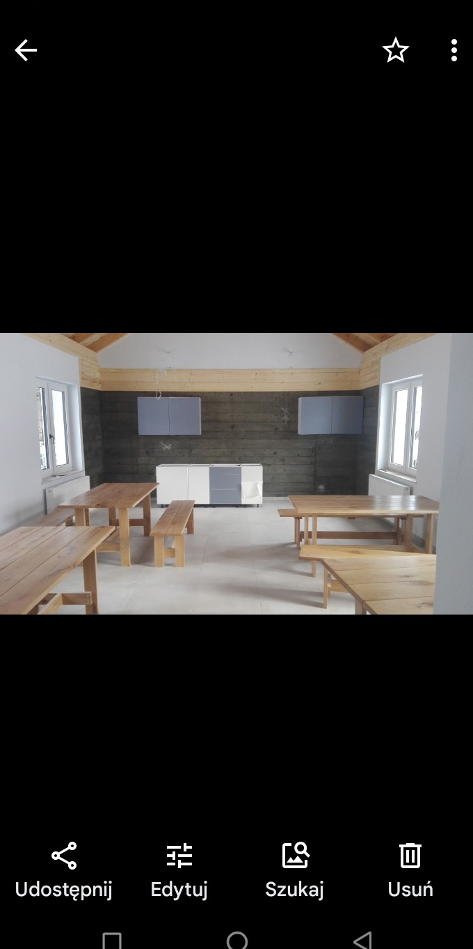 Surowe drewno na ścianie, jasne panele podłogowe, drewniane stoły i ławy w nowym pomieszczeniu z białymi ścianami i oknami, szare szafki wiszące i biało-szara komoda.