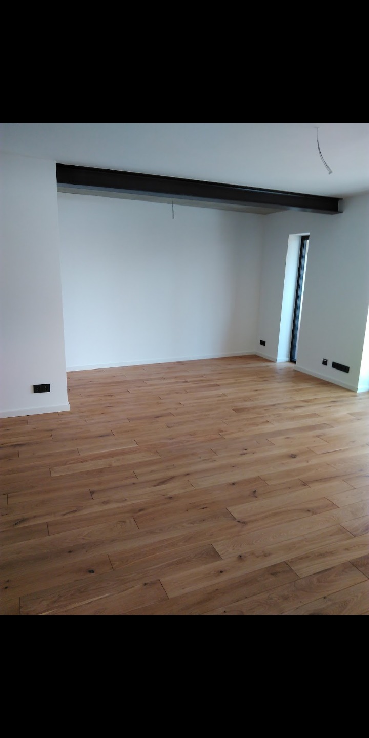 Jasne panele podłogowe ułożone w przestronnym, minimalistycznym pokoju z białymi ścianami i czarną belką stropową, widoczne gniazdka elektryczne.