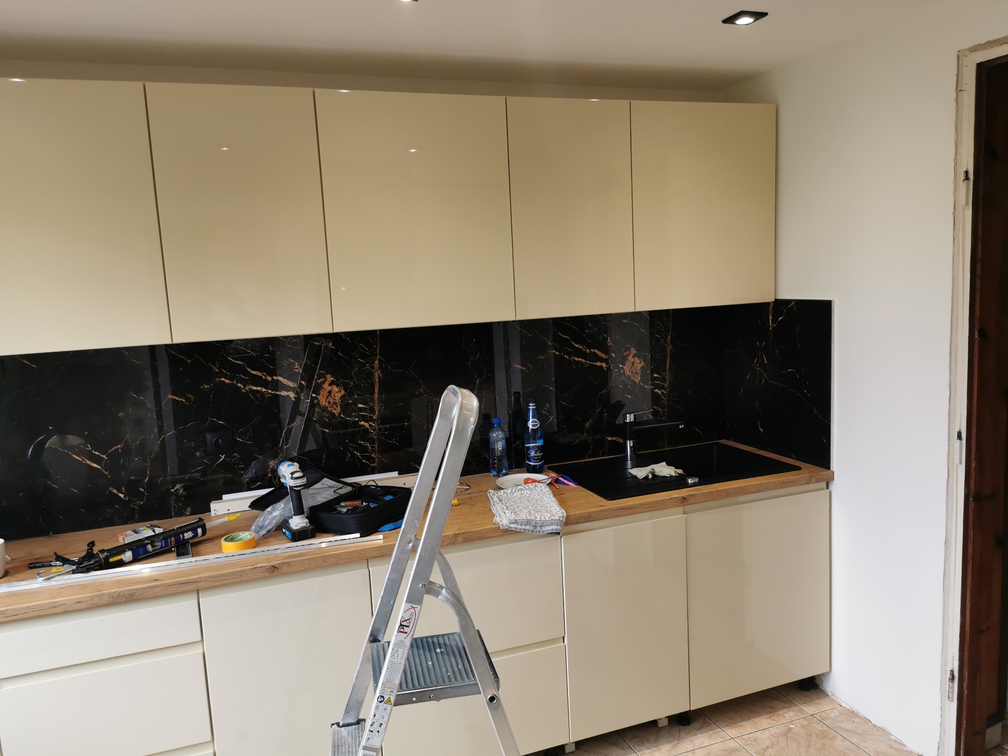 Montaż paneli szklanych z motywem czarnego marmuru nad blatem w kuchni, widoczna drabina, narzędzia i akcesoria montażowe na blacie.