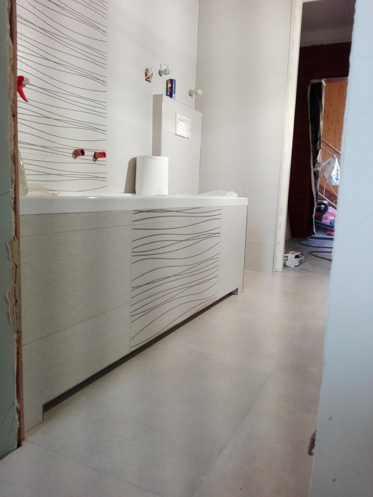 Niedokończona łazienka z białymi płytkami z szarym wzorem, widoczne wystające rury i niedokończone wykończenia oraz odkurzacz w przejściu do kolejnego pomieszczenia.