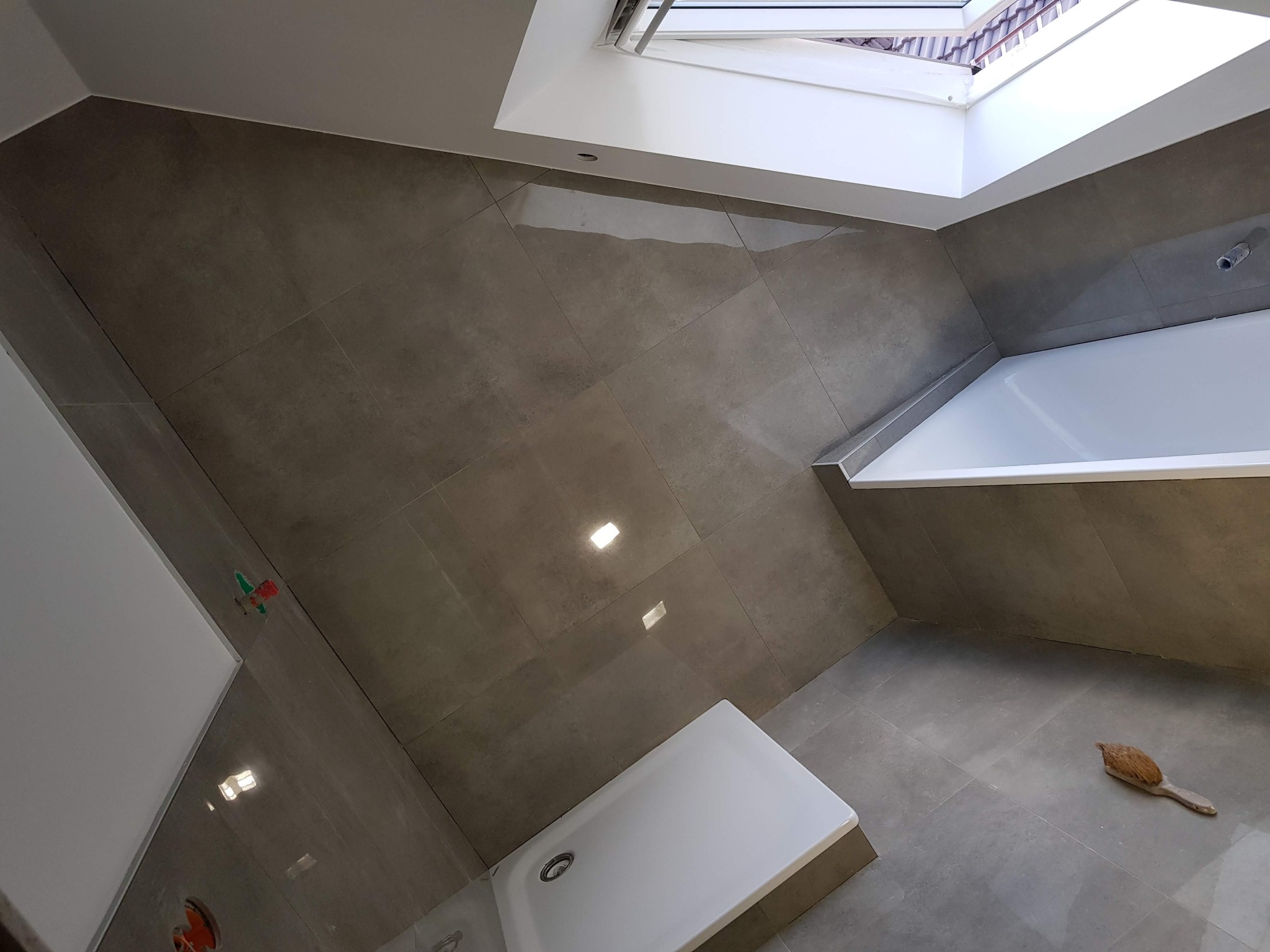 Wykończona łazienka z szarymi, matowymi płytkami, białą wanną i brodzikiem oraz oknem dachowym, widoczna szczotka do czyszczenia fug na podłodze.