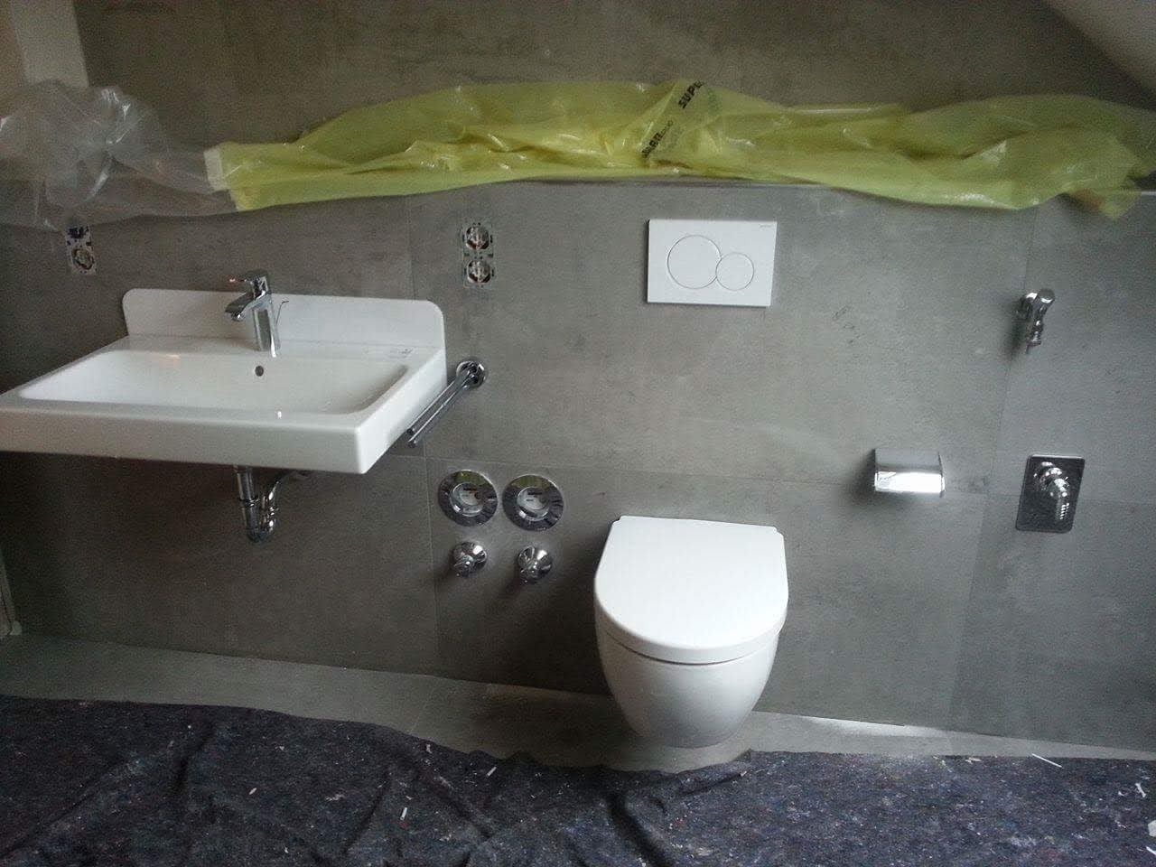 Montaż umywalki i toalety w łazience z szarą okładziną, zabezpieczona podłoga, widoczne przygotowane instalacje.