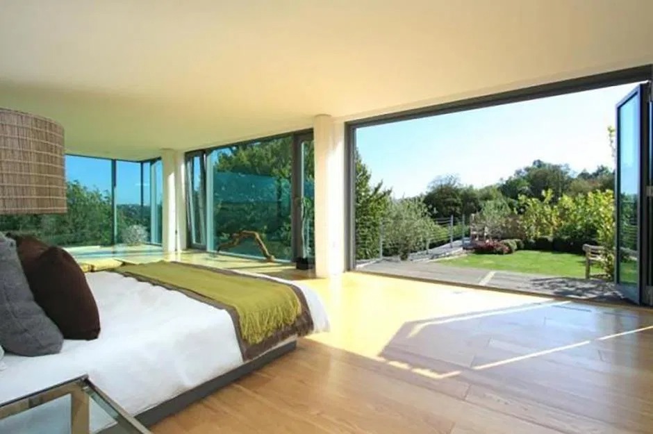 Jasna sypialnia z panoramicznymi oknami wychodzącymi na ogród, drewniana podłoga i nowoczesne meble.