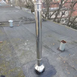 Błyszczący, metalowy komin na płaskim, pokrytym papą dachu, z widocznymi śladami smoły wokół podstawy, w tle drzewa bez liści i zabudowania.