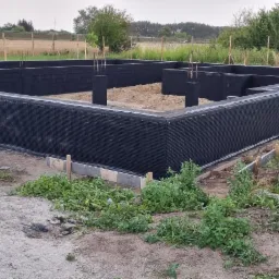 Wykopane i zabezpieczone fundamenty domu jednorodzinnego z widocznym zbrojeniem pionowym, otoczone ziemią i zielenią, w tle pole i droga.