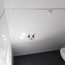 Damian Szymański - Nowoczesna łazienka z białymi płytkami na ścianach i suficie, ciemnoszarą podłogą i widocznymi przyłączami wodnymi do baterii umywalkowej.