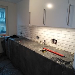 Damian Szymański - Białe, prostokątne płytki ułożone na ścianie w kuchni, widoczna instalacja wodna, blat zabezpieczony folią, poziomica i miarka leżą na blacie, białe szafki wiszące z długimi, srebrnymi uchwytami.