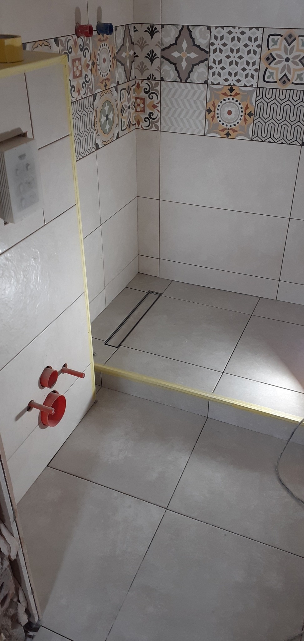 Fragment łazienki w trakcie wykańczania: ułożone jasnoszare płytki podłogowe i ścienne, dekoracyjny pas z wzorzystych płytek, widoczny odpływ liniowy, przygotowane przyłącza hydrauliczne i żółta...