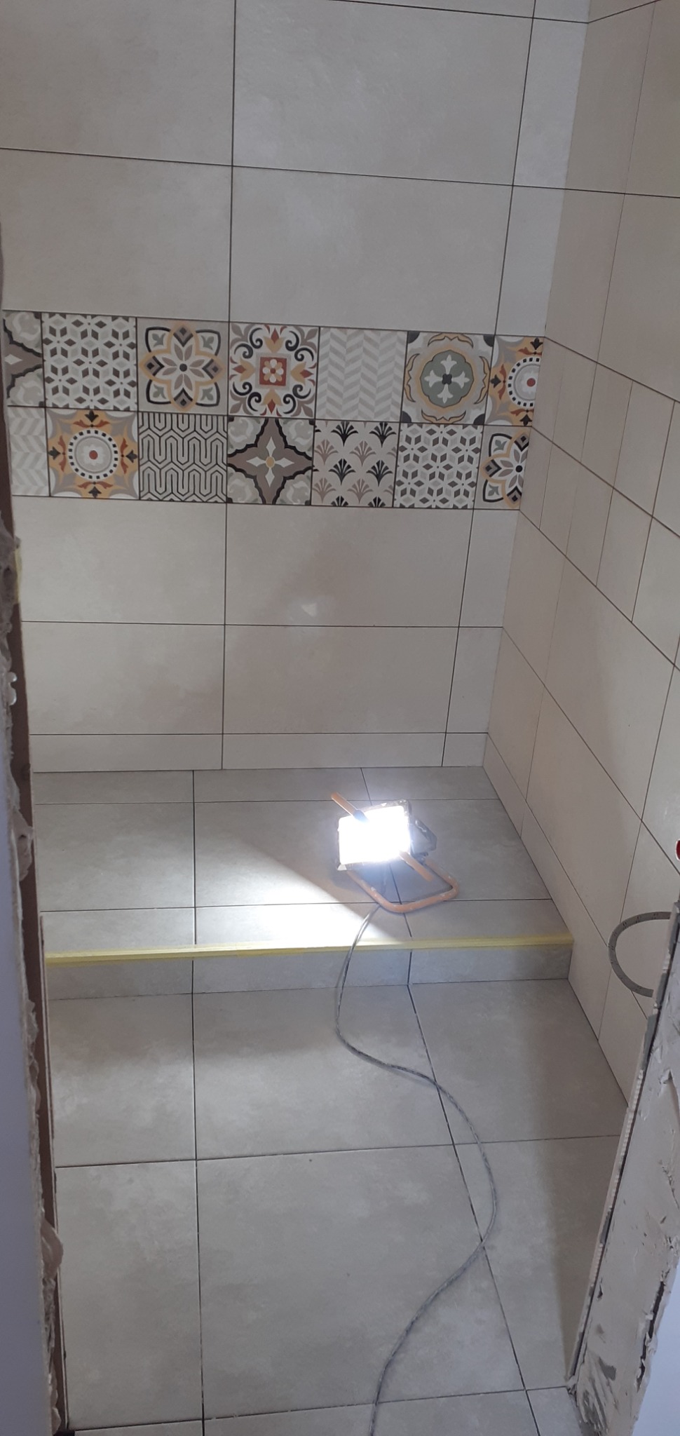 Fragment łazienki w trakcie prac wykończeniowych: świeżo położone płytki podłogowe i ścienne z dekoracyjnym pasem wzorzystych płytek, oświetlone lampą roboczą na podłodze, z widocznymi śladami po...