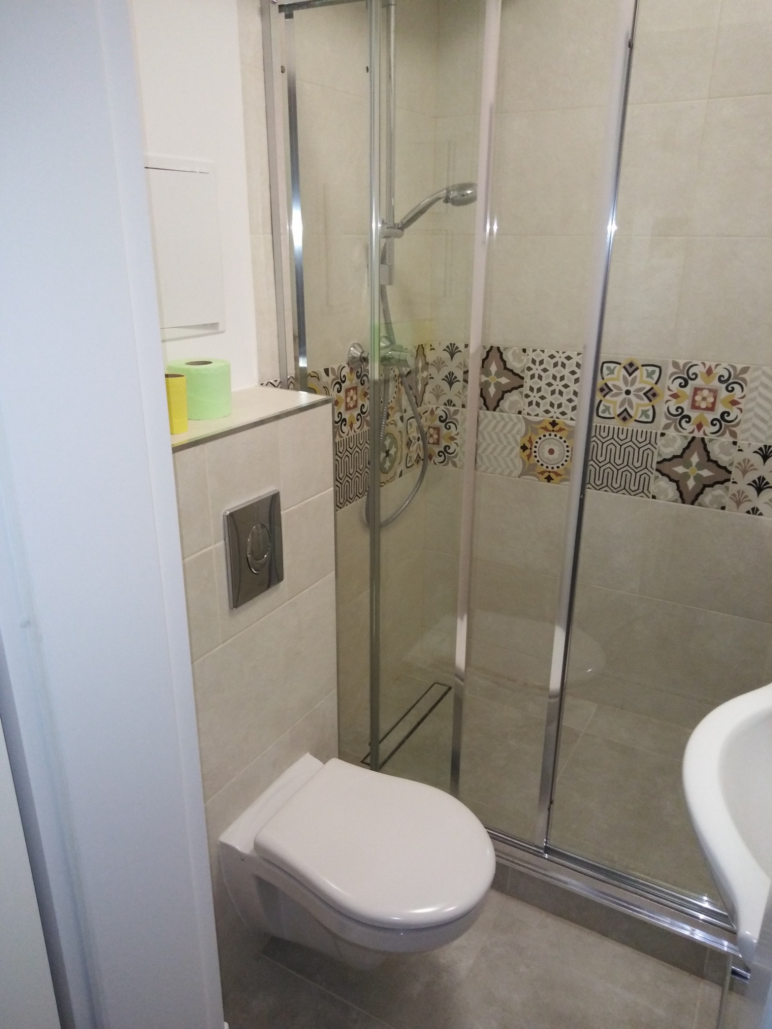 Kompaktowa łazienka z kabiną prysznicową za szklanymi drzwiami, podwieszana toaleta z chromowanym przyciskiem spłukiwania i dekoracyjne płytki nad prysznicem.