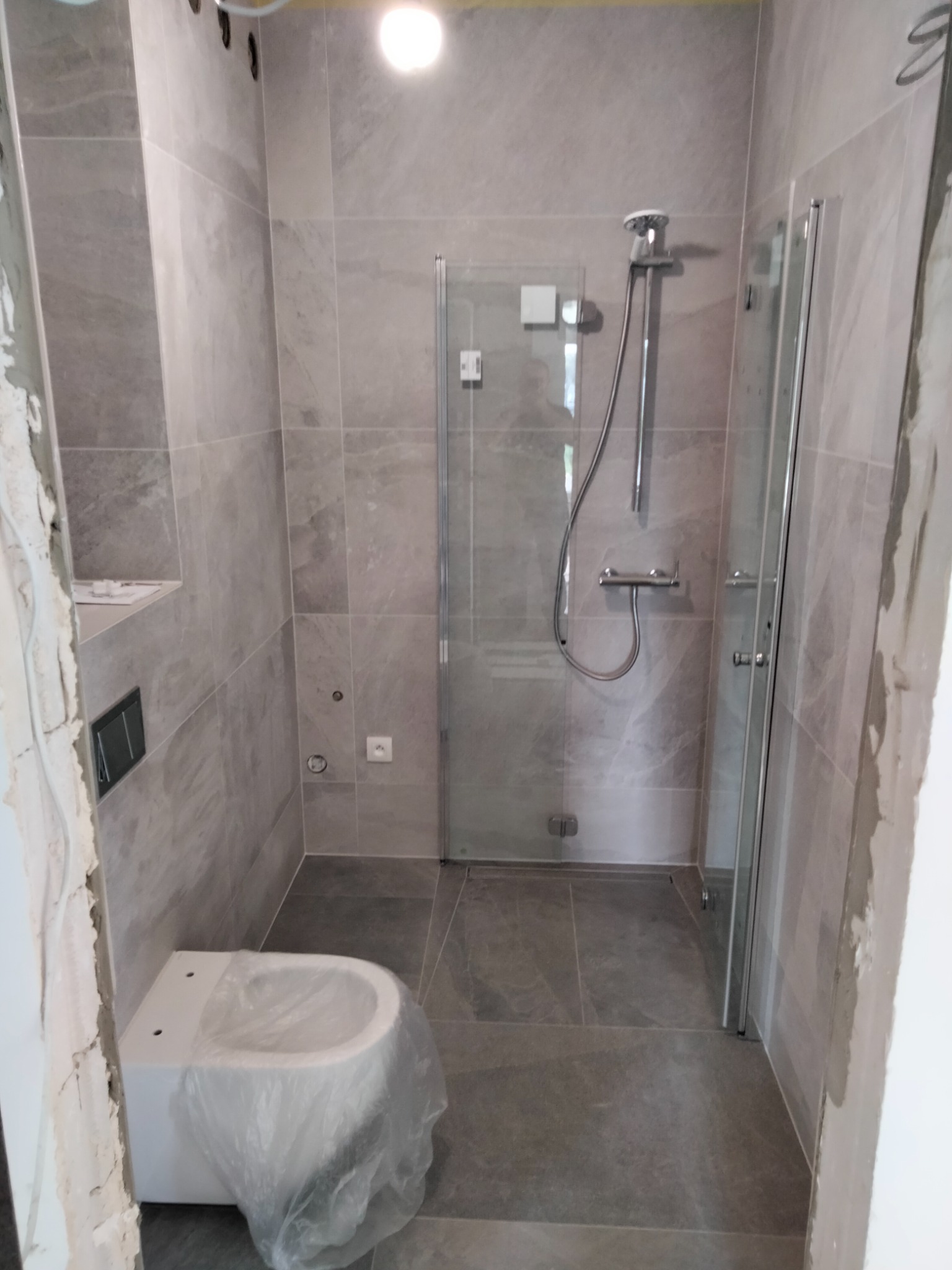 Szara łazienka w trakcie remontu, widoczna kabina prysznicowa z deszczownicą, podwieszana toaleta zabezpieczona folią, szare płytki na ścianach i podłodze, surowy stan wykończenia przy futrynie.